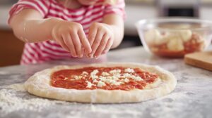 Child sprinkling cheese onto pizza dough covered with tomato sauce on a floured countertop