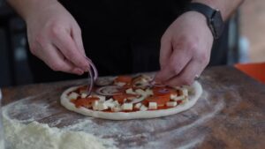 Hands Adding Sliced Red Onions to A Pizza with Sauce, Cheese, Pepperoni, and Mushrooms on A Floured Wooden Surface