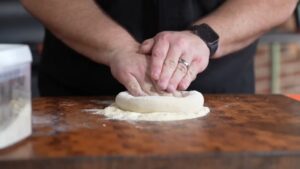 Hands Pressing Down on A Ball of Pizza Dough on A Floured Wooden Surface