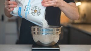 A Person Pouring Gluten-Free Flour from A King Arthur Bag Into a Stainless Steel Mixing Bowl on A Digital Scale