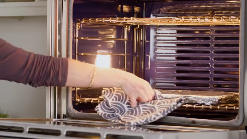 A Person Using an Oven Mitt to Take a Foil-Covered Tray out Of a Hot Oven