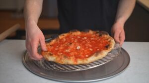 A Person Holding a Freshly Baked Gluten-Free Pizza with A Charred Crust, Topped with Tomato Sauce and Garlic, on A Cooling Rack