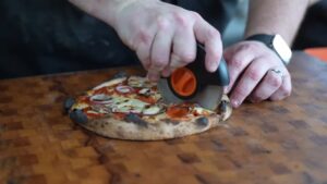 A Freshly Cooked Pizza Is Being Sliced on A Wooden Cutting Board Using a Pizza Cutter